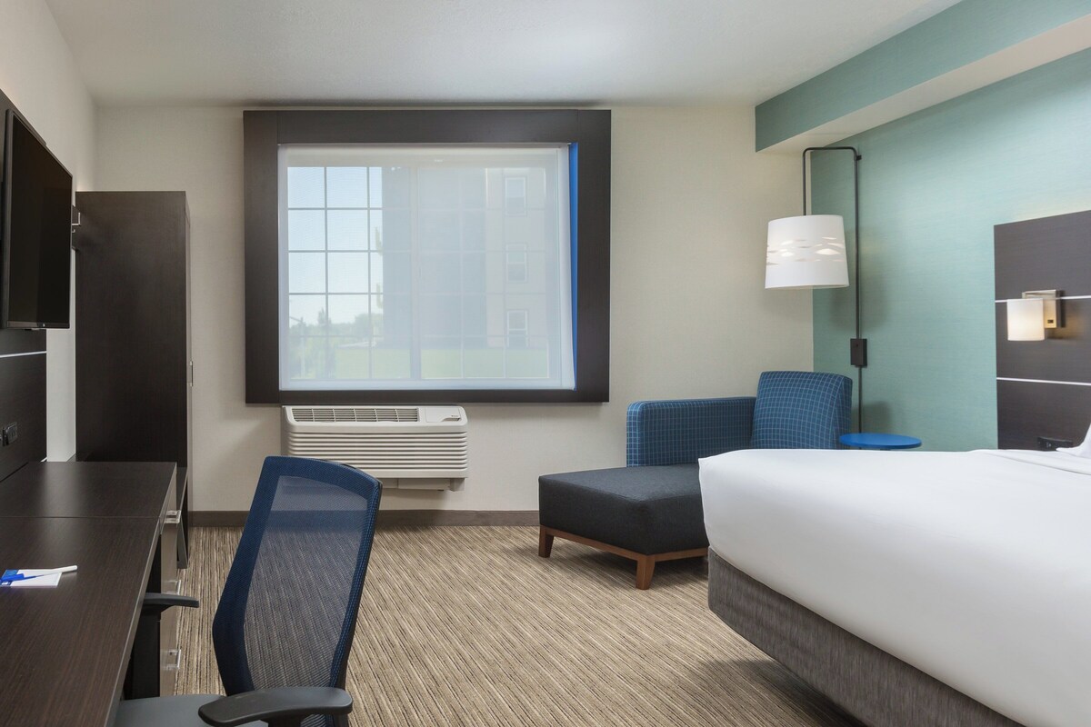 A hotel room with a bed, a desk, a chair, a window, and a wall-mounted lamp.