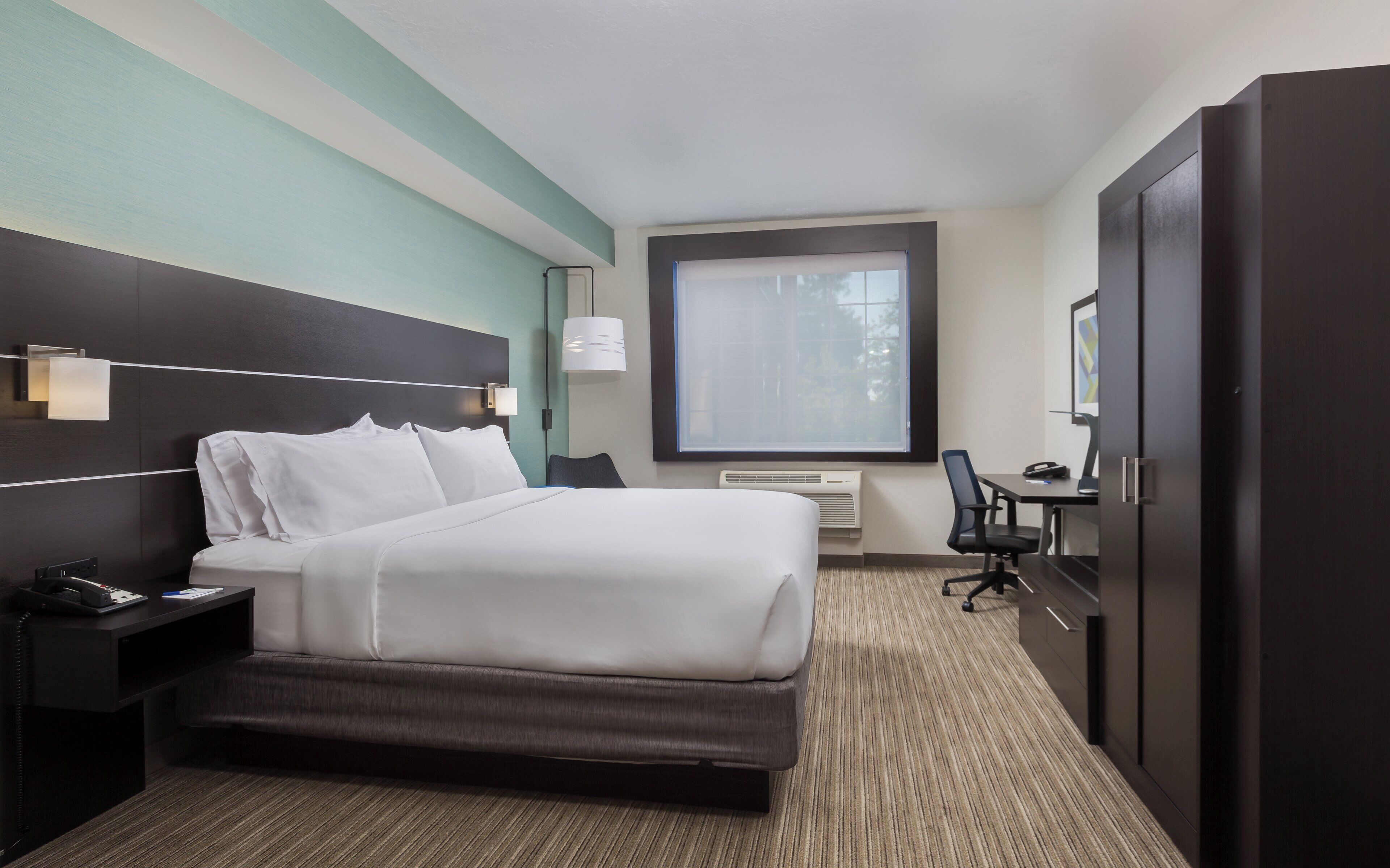 A hotel room with a bed, bedside table, desk, chair, and a large window.