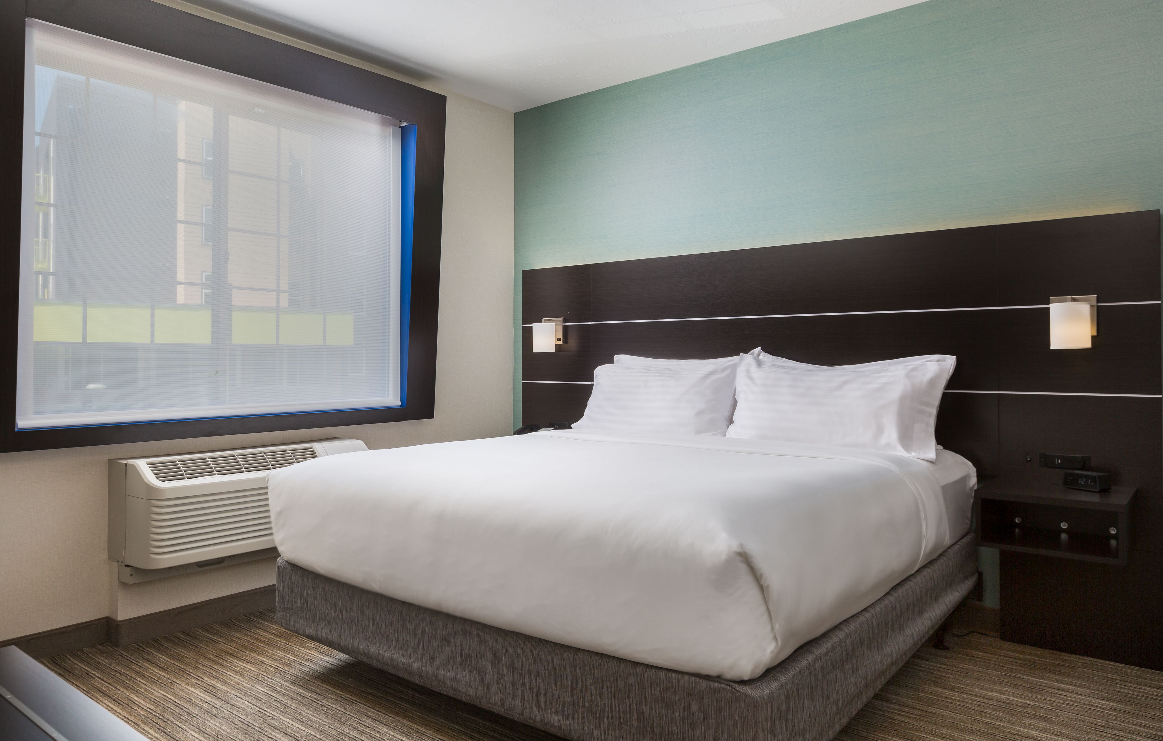 Habitación de hotel con una cama grande, una ventana con persianas, un aire acondicionado de pared y una mesita de noche con lámpara.