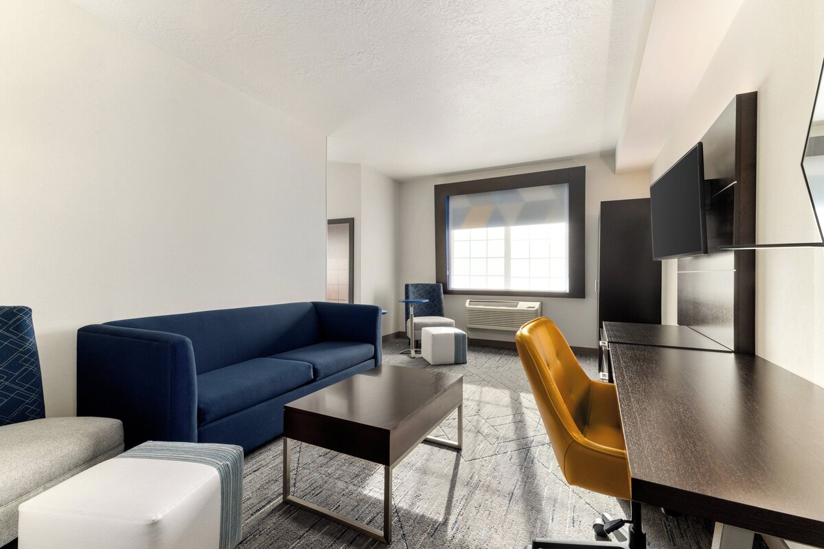 A modern hotel room with a blue sofa, a small white ottoman, a dark wooden table, a yellow chair, and a large window providing natural light.