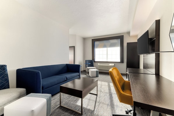 Un hotel moderno con un sofá azul, un pequeño reposapiés blanco, una mesa de madera oscura, una silla amarilla y un gran ventanal que proporciona luz natural.