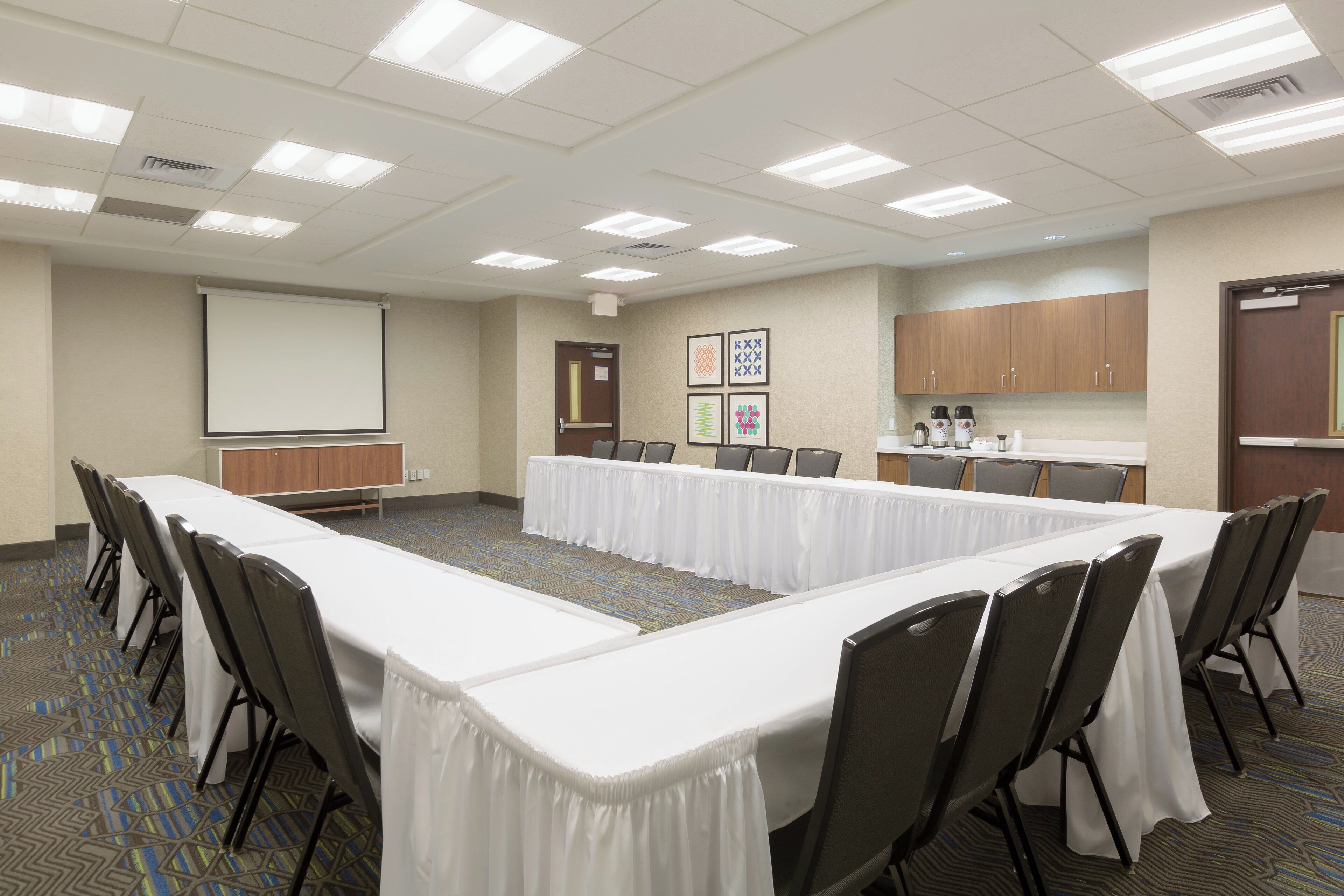 A conference room with a long table, chairs, a projector screen, and a kitchenette area.