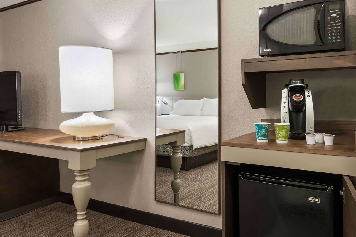 A hotel room with a bed, a mirror, a microwave, a coffee maker, and a refrigerator.