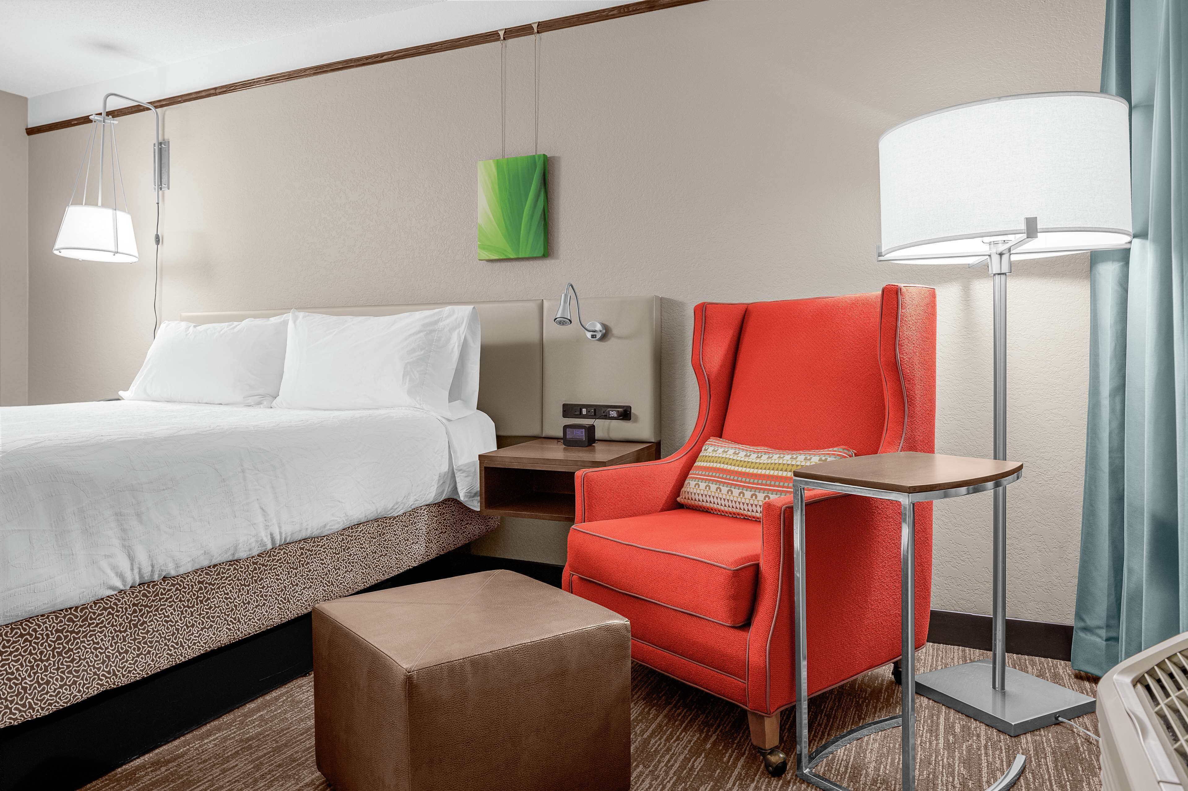 A hotel room with a bed, a red armchair, a small table, a nightstand, a lamp, and a green artwork on the wall.