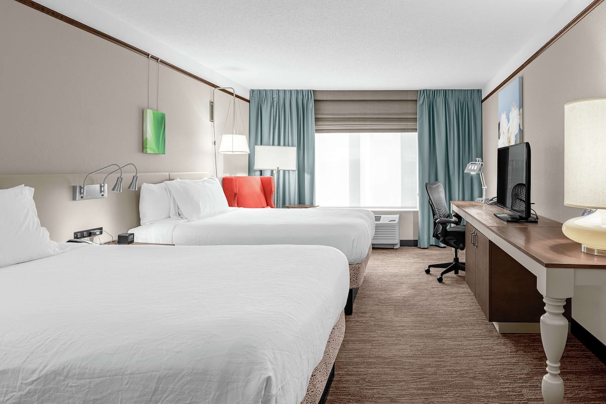 A hotel room with two beds, a desk, a chair, a TV, and a window with curtains.