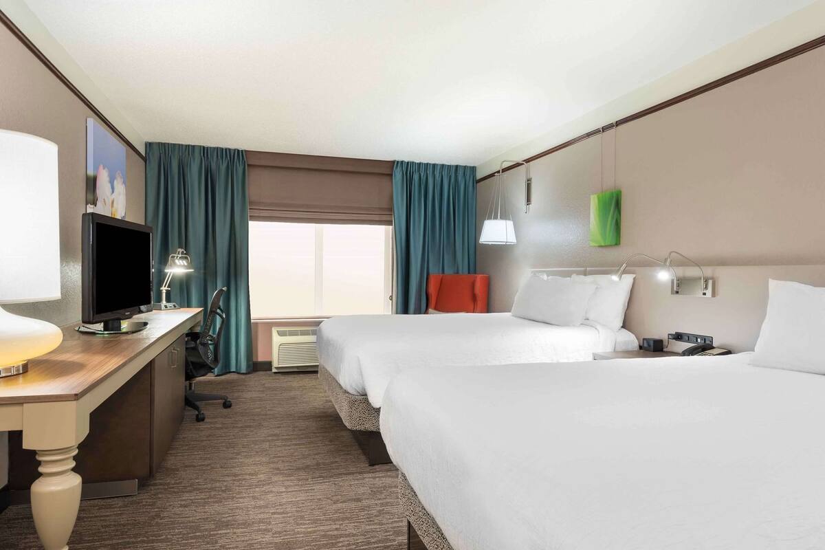 A hotel room with two beds, a desk with a computer, a TV, a lamp, and a window with curtains.
