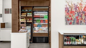 Ein Hotel-Rezeptionsbereich mit einem Getränkekühlschrank, der mit verschiedenen Getränken gefüllt ist, einer kleinen Auswahl an Snacks und einem Bild an der Wand.