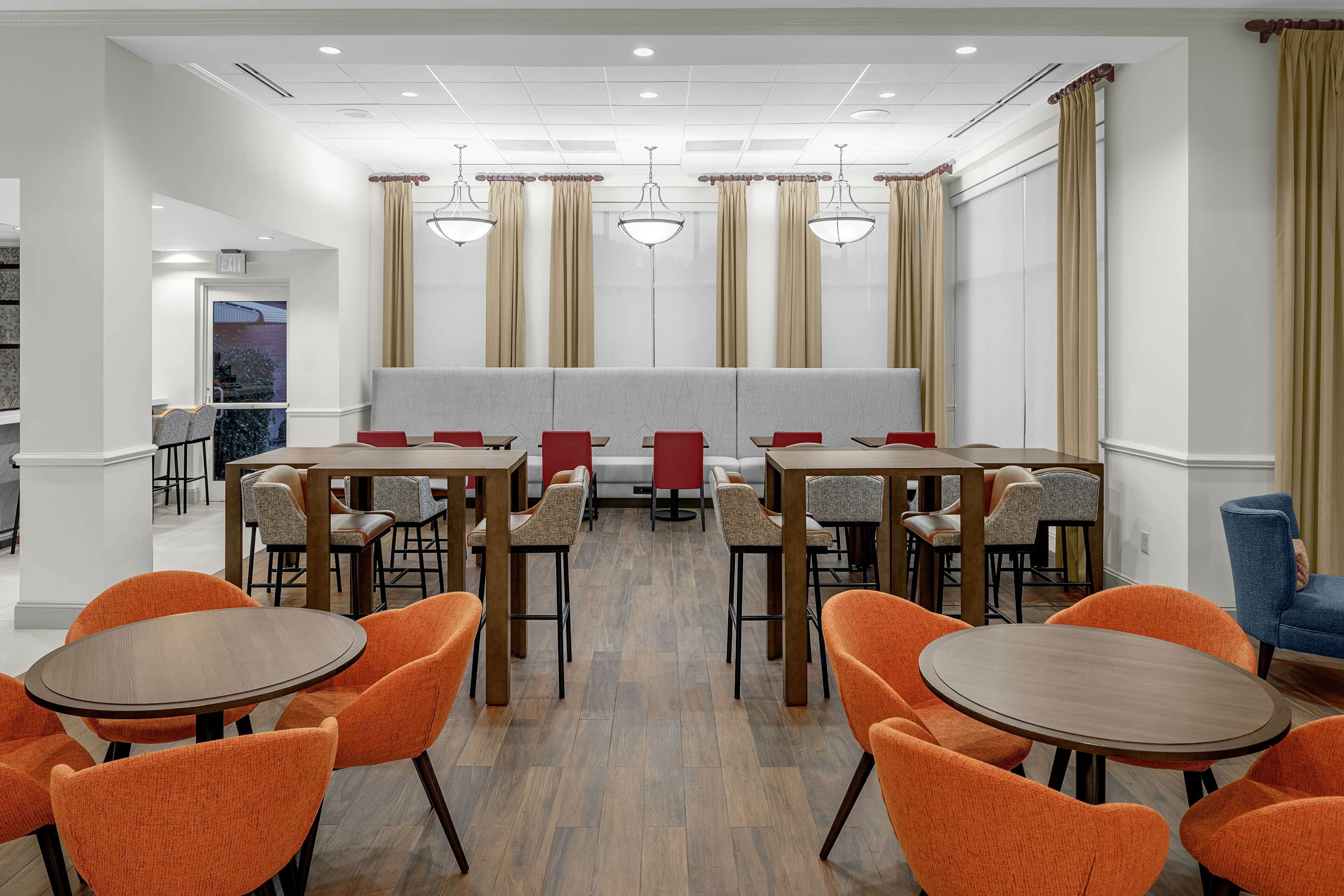 Un espace de restauration d’hôtel avec des tables rondes et des chaises orange, un banc rembourré gris et des rideaux beige.
