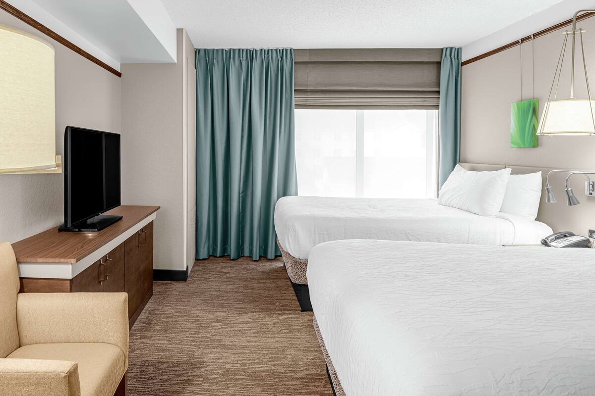 A hotel room with two beds, a TV on a wooden cabinet, a desk, and a window with curtains.