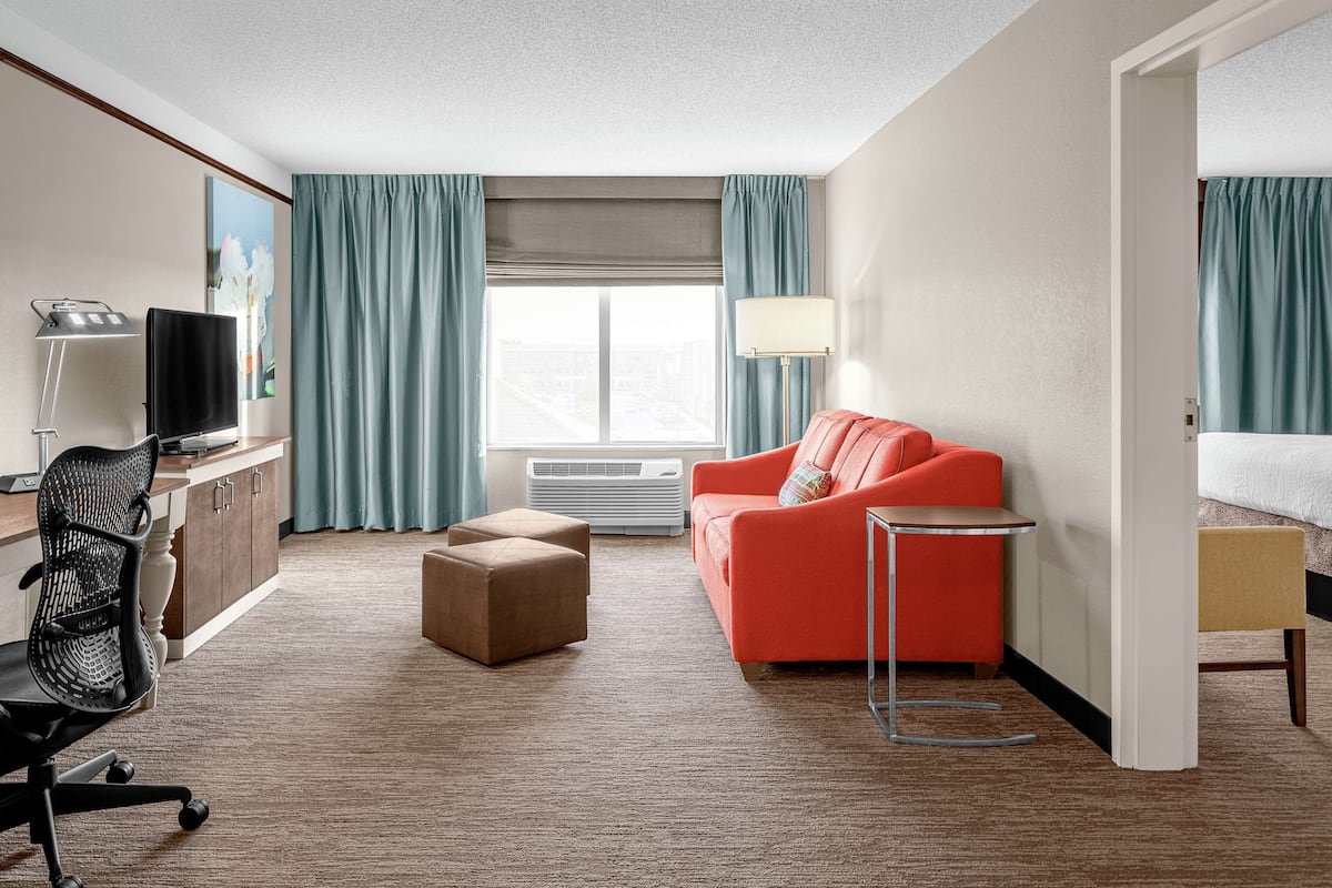 A hotel room with a desk, chair, red sofa, ottoman, lamp, and a bed.