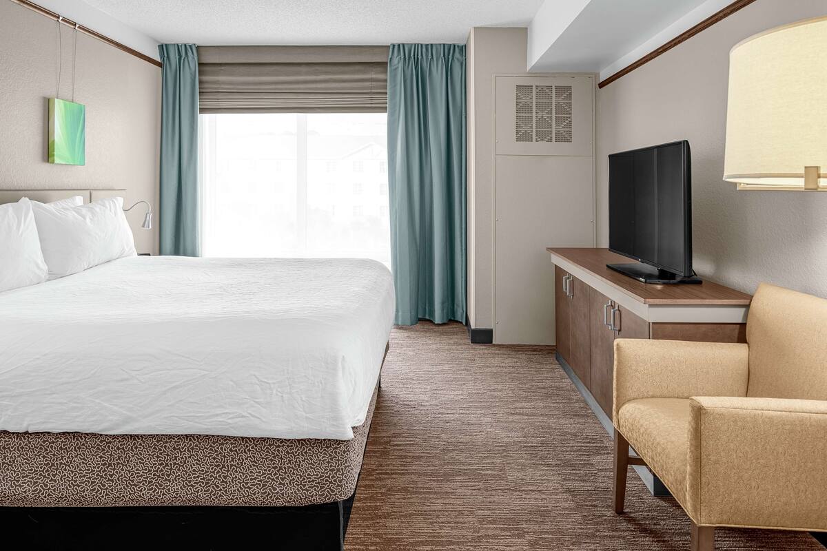 A hotel room with a large bed, a television on a wooden cabinet, a beige armchair, and a window with curtains.