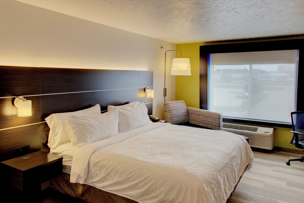 Una habitación de hotel con cama, mesitas de noche, una silla, una ventana y una lámpara de pared.