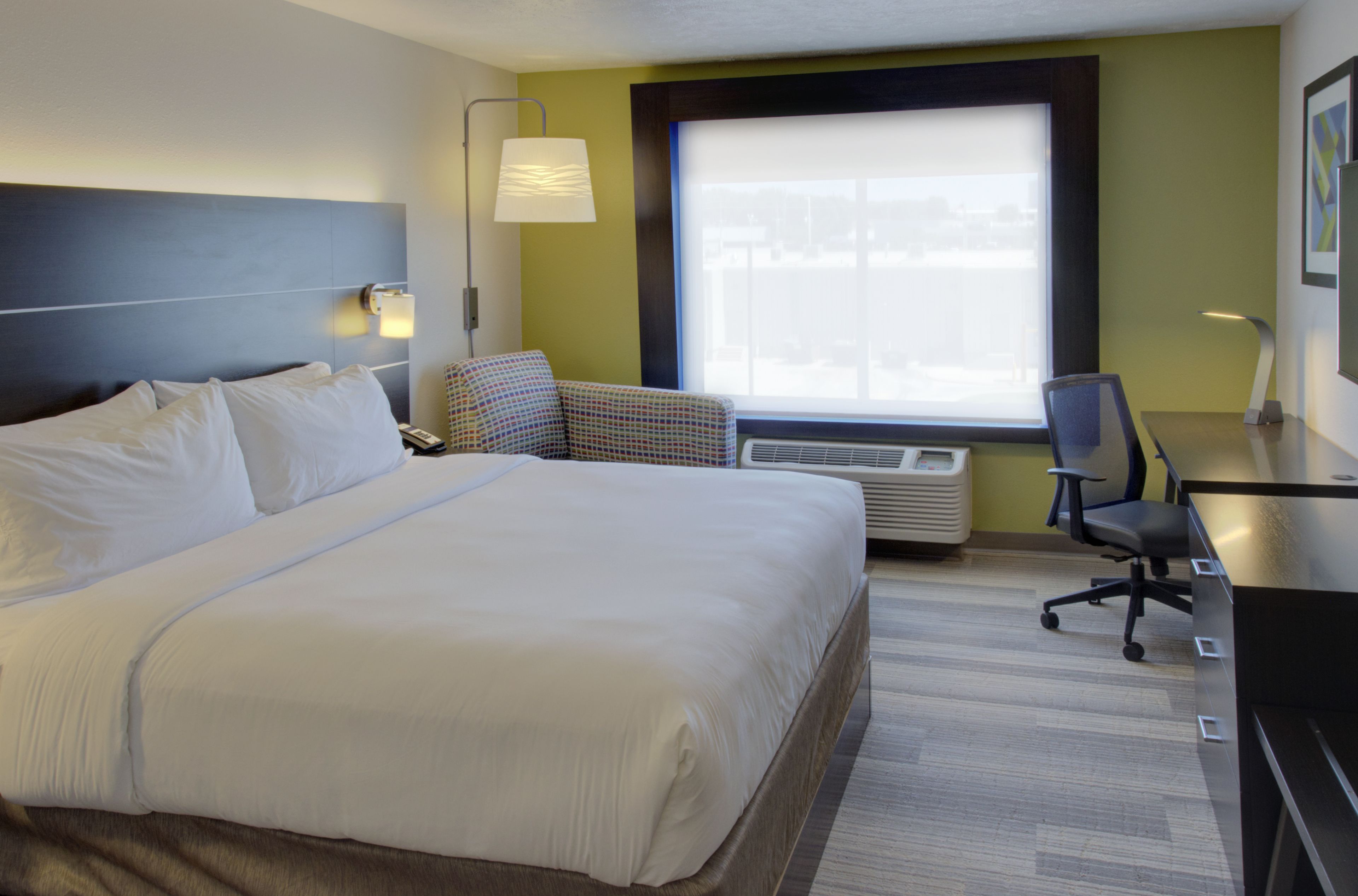 A hotel room with a large bed, a desk, a chair, a lamp, and a window with blinds.