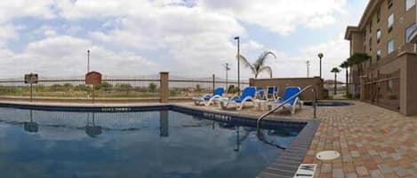 A swimming pool with lounge chairs, a fenced area, and a building in the background.