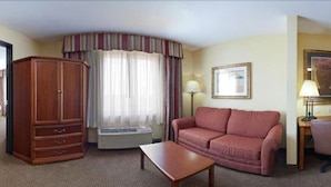A hotel room with a wooden wardrobe, a pink sofa, a wooden coffee table, a window with curtains, a lamp, and a framed picture on the wall. - Holiday Inn Express Hotel & Suites Laredo-Event Center Area by IHG (Laredo)