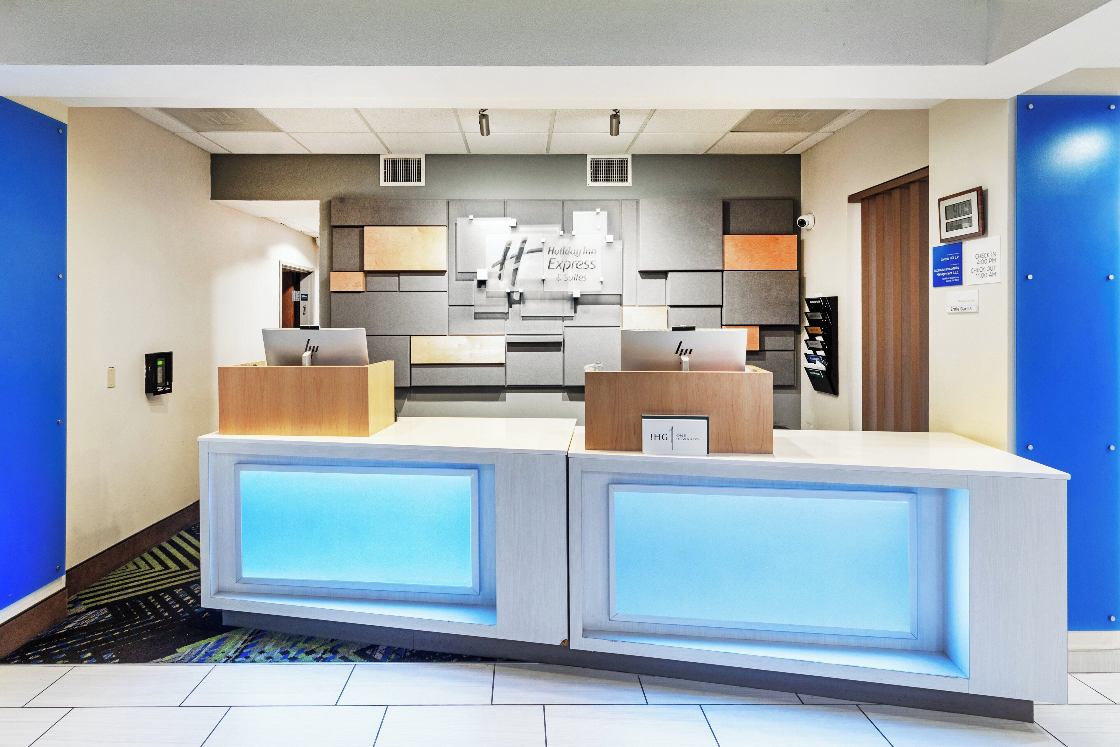 A reception desk with a modern design, featuring a flat-screen monitor, a laptop, and a sign indicating the hotel name.