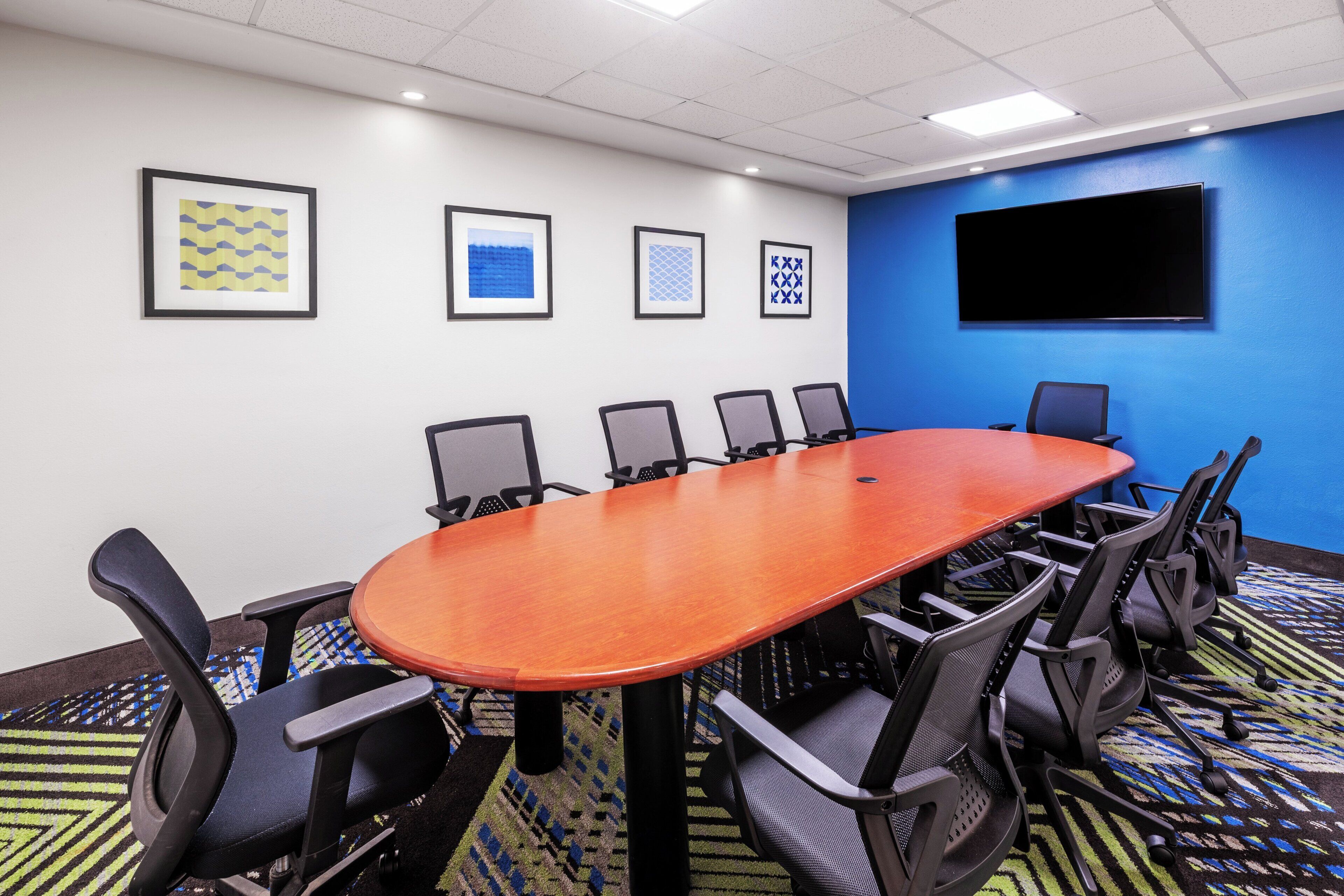 A conference room with a long table and six chairs, a flat-screen TV on the wall, and framed artwork on the wall.