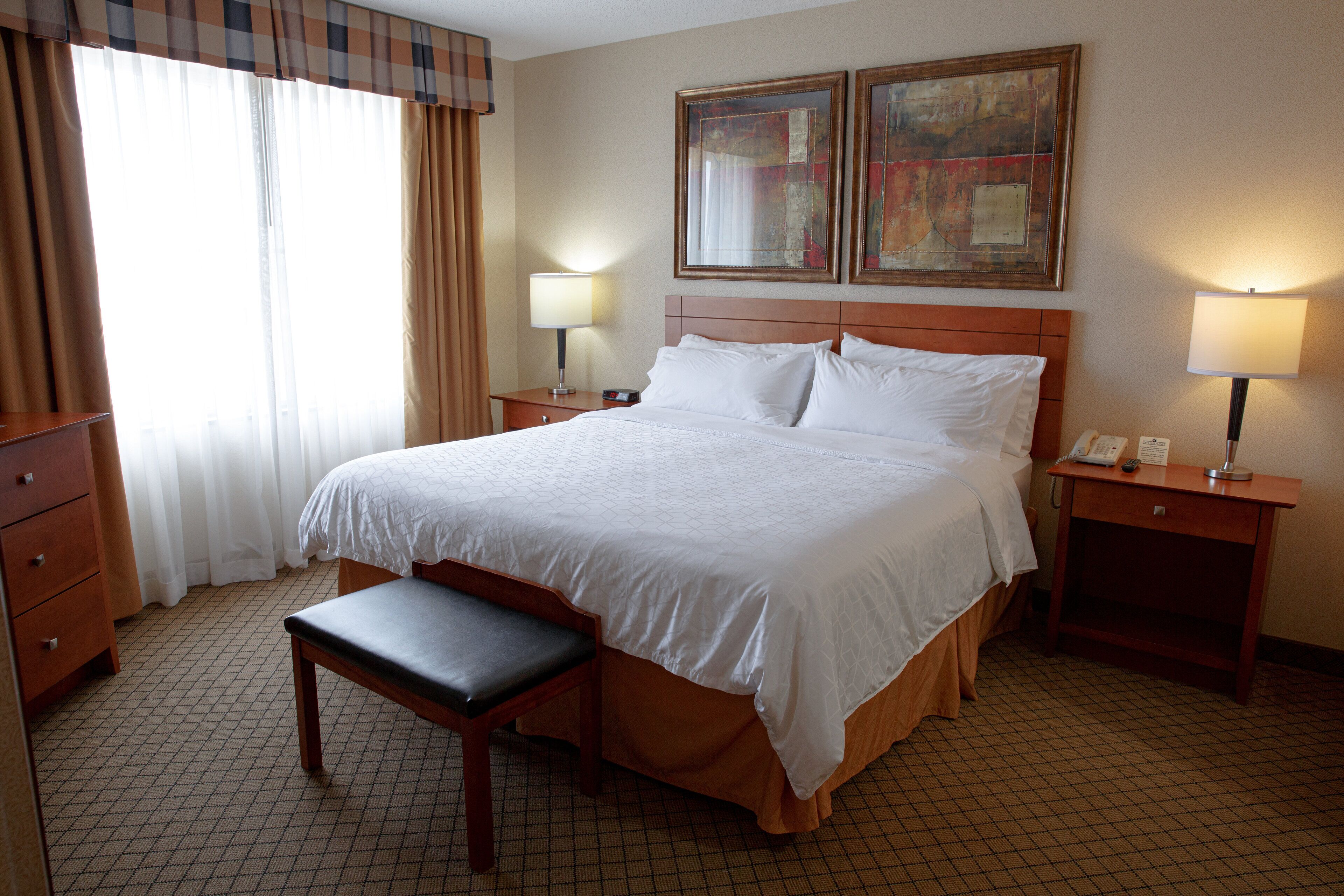 A hotel room with a large bed, a wooden bench, two bedside tables with lamps, a dresser, and a window with curtains.