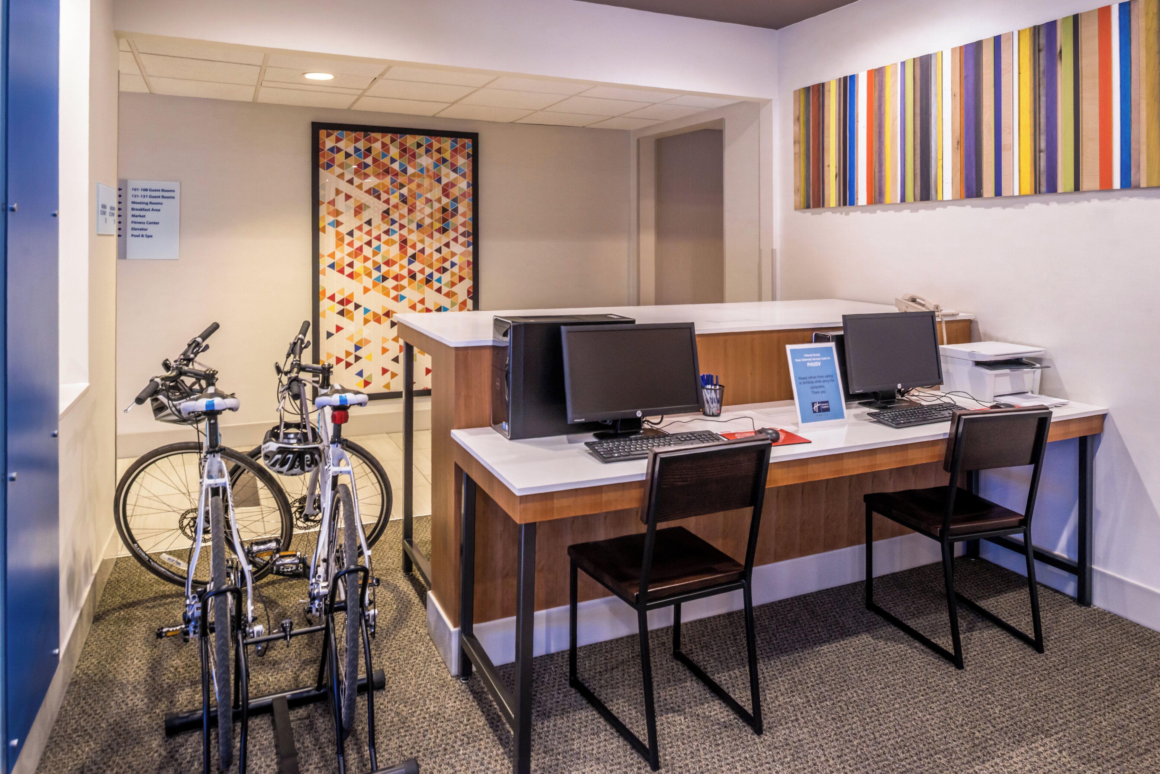 A hotel reception area with two computer workstations, a bicycle rack, and a colorful geometric wall art.