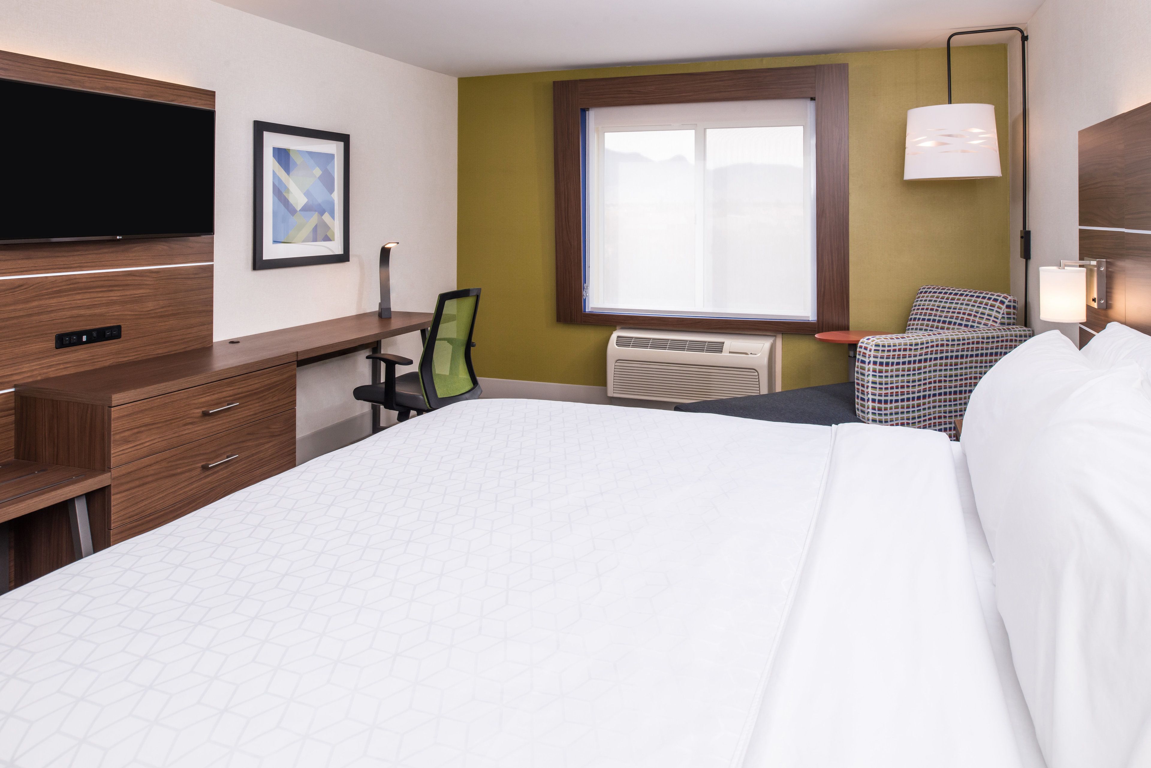 A hotel room with a large bed, a desk with a chair, a flat-screen TV, a window with a view, and a lamp.