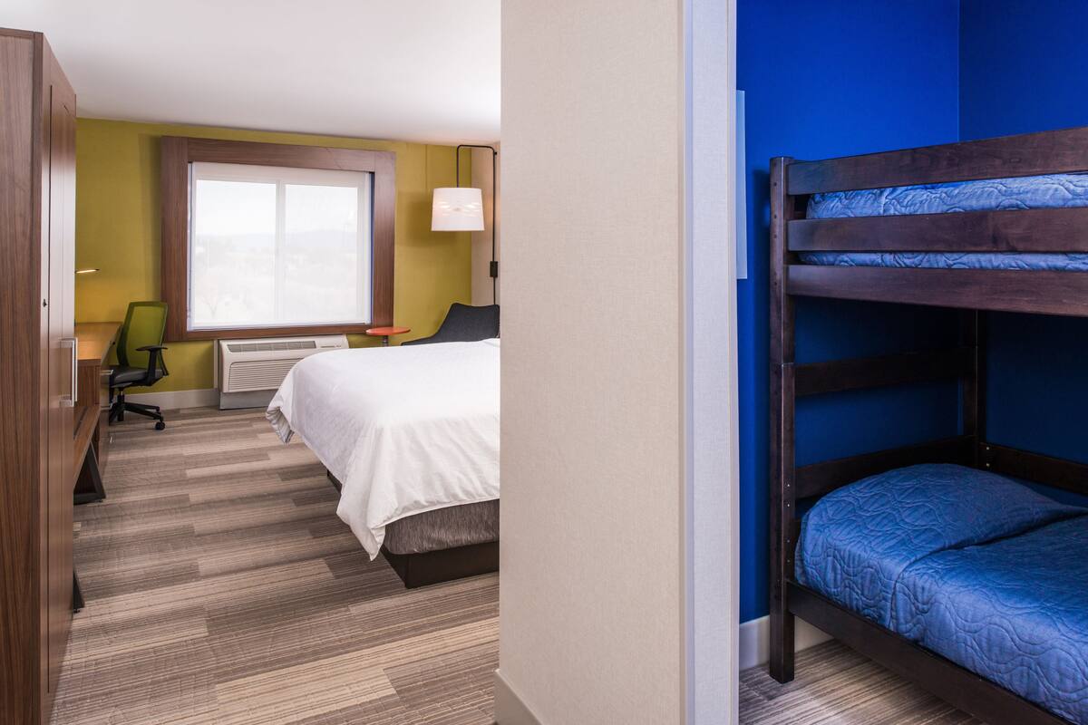 A hotel room with a bunk bed, a desk with a chair, a window with blinds, and a lamp.