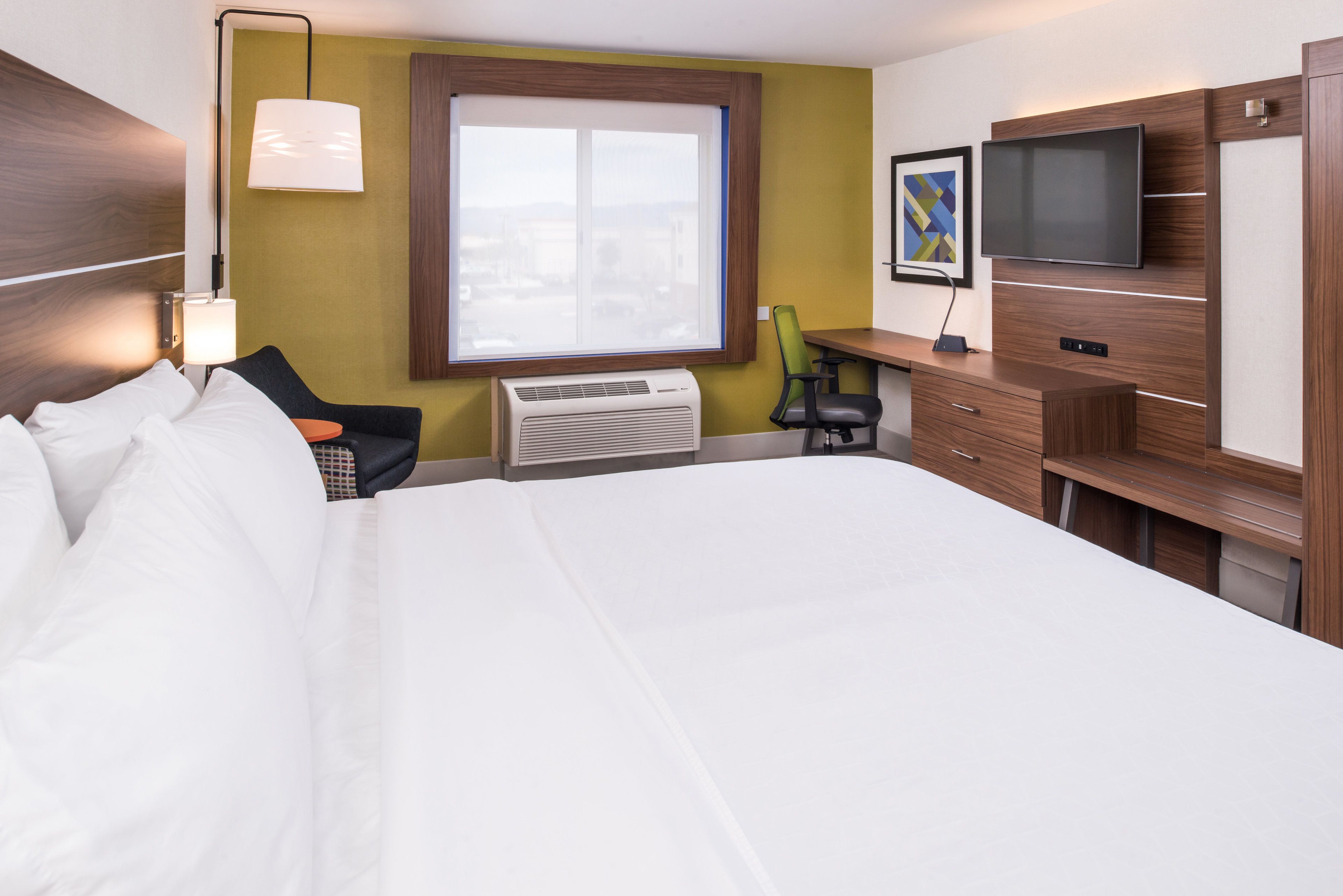 A hotel room with a large bed, a desk with a chair, a flat-screen TV, and a window with a view of buildings.