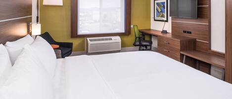 A hotel room with a large bed, a desk with a chair, a flat-screen TV, and a window with a view of buildings.