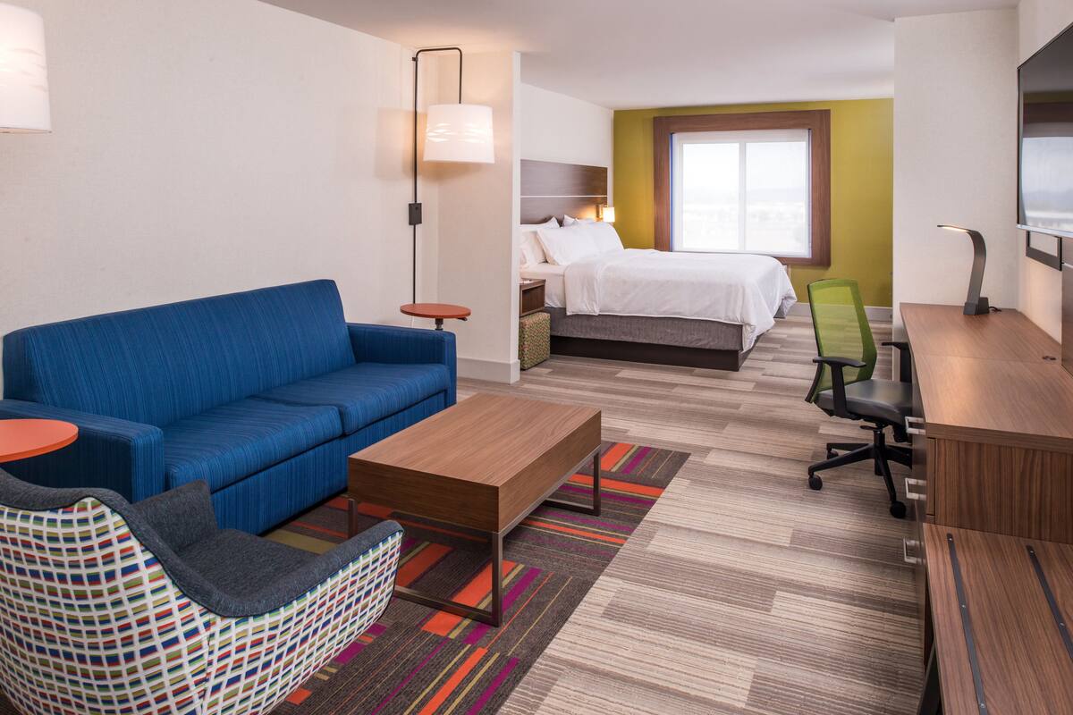 A hotel room with a blue sofa, a wooden coffee table, a green office chair, a bed with white bedding, and a TV mounted on the wall.