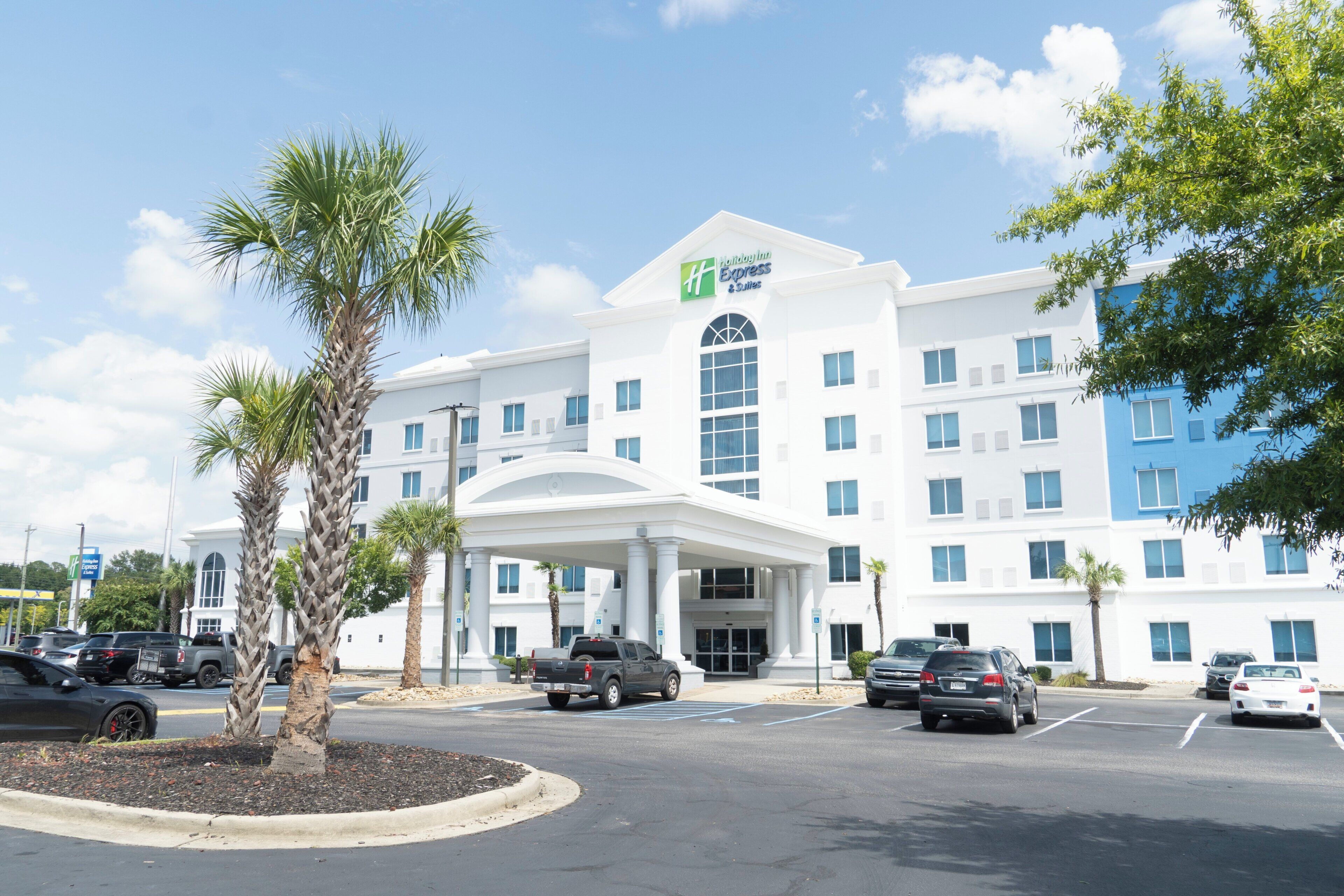 A Holiday Inn Express & Suites hotel with a central entrance, parking lot, and palm trees.