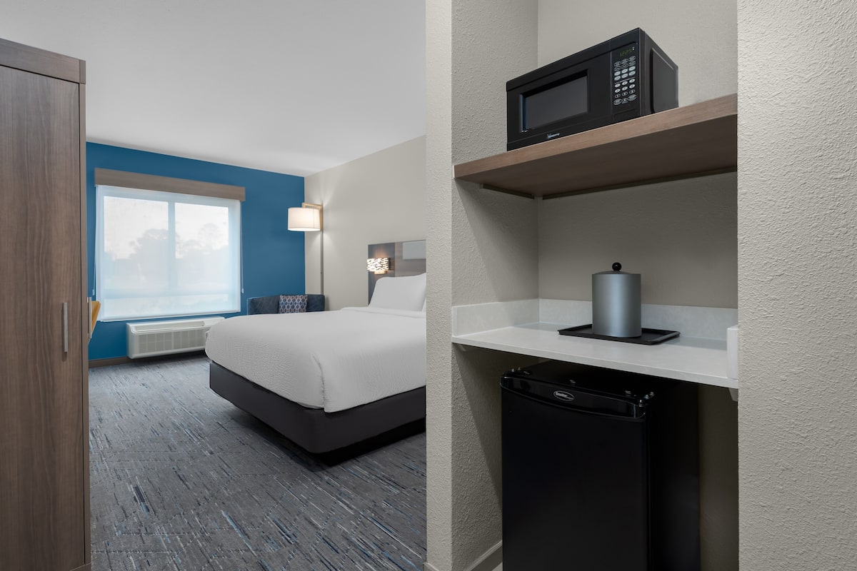A hotel room with a bed, a microwave, a small refrigerator, and a window with blinds.
