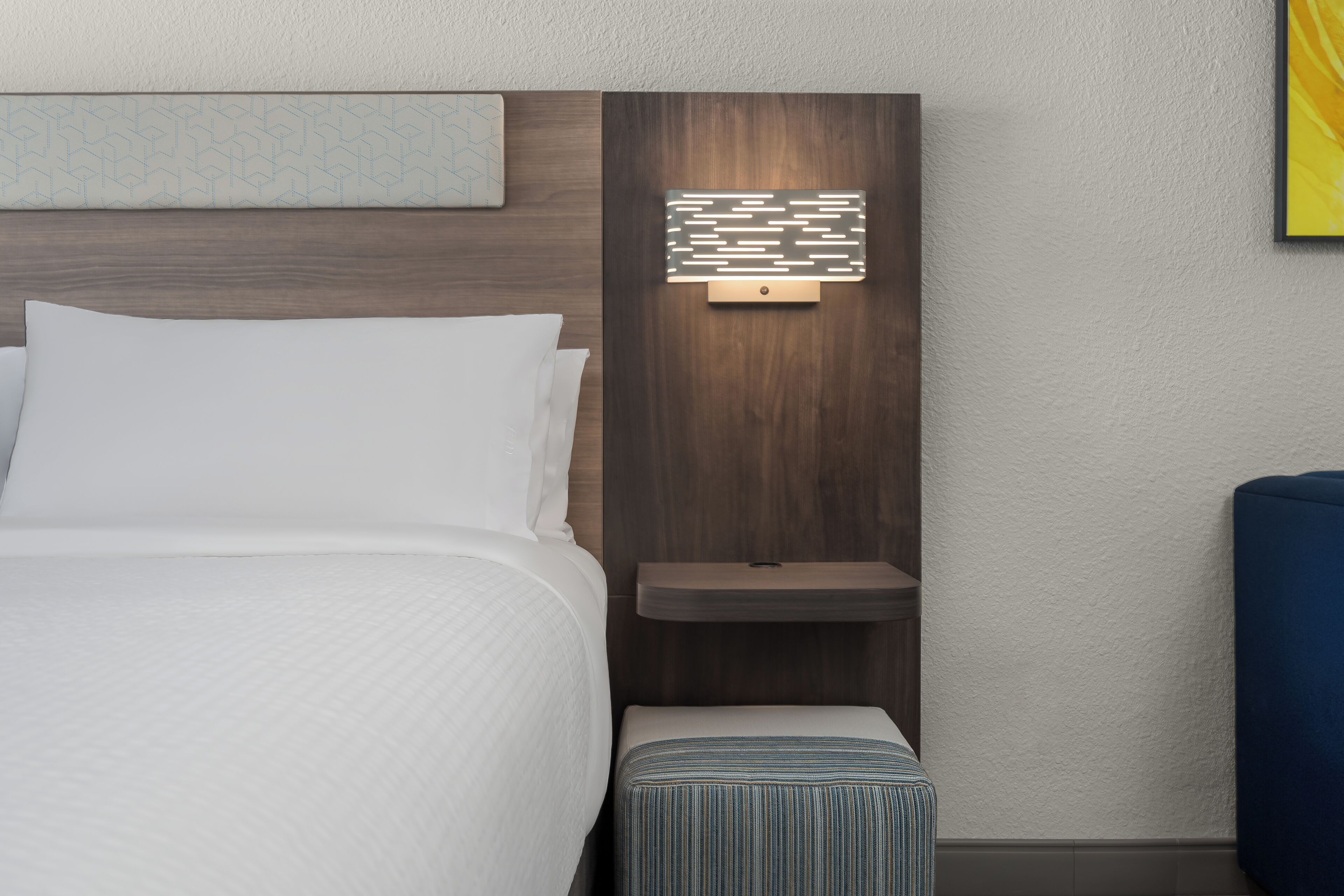 A hotel room with a bed, a bedside table, a nightstand, and a wall-mounted light fixture.