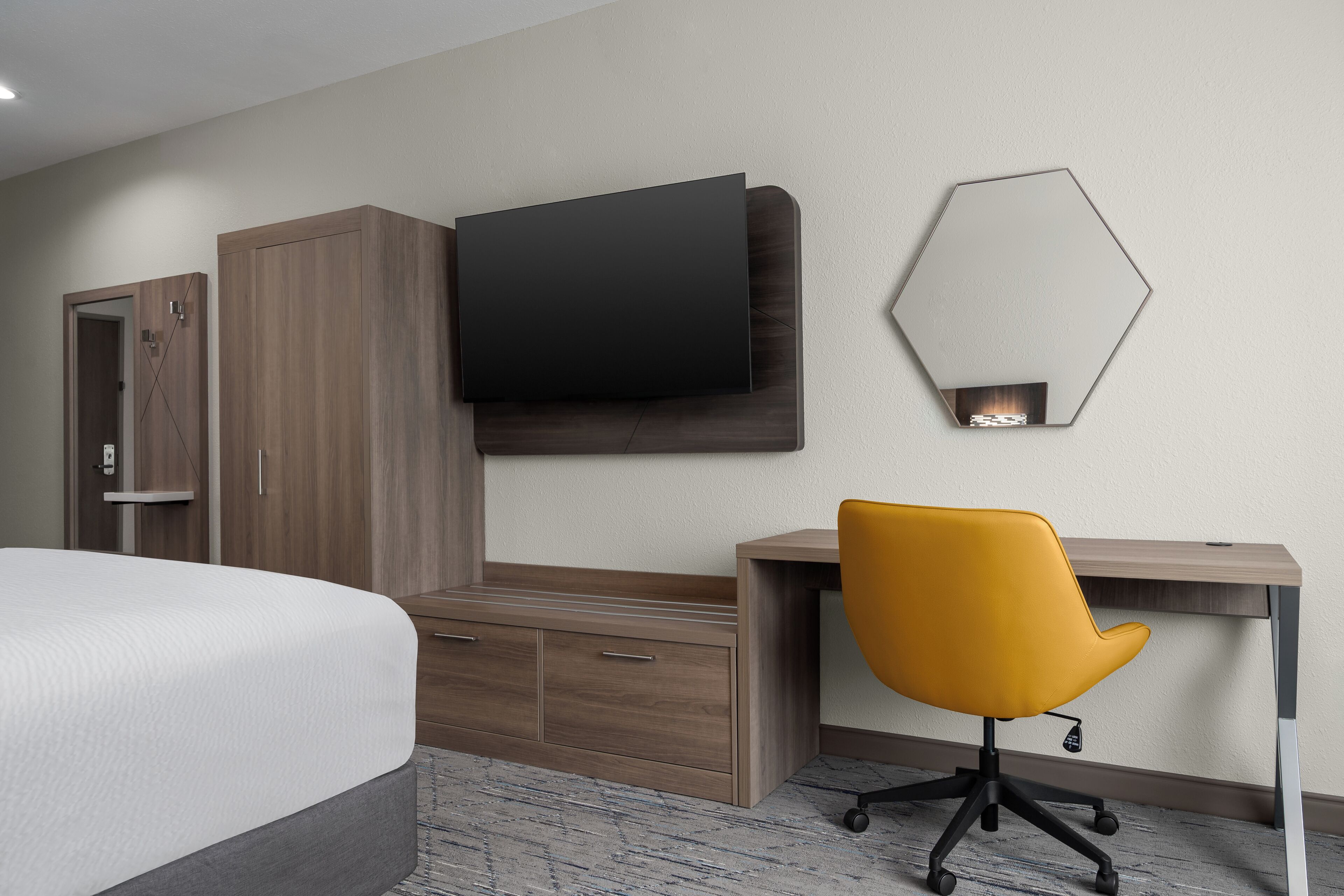 Quarto de hotel com cama, escrivaninha, cadeira, televisão na parede, guarda-roupa e espelho hexagonal.