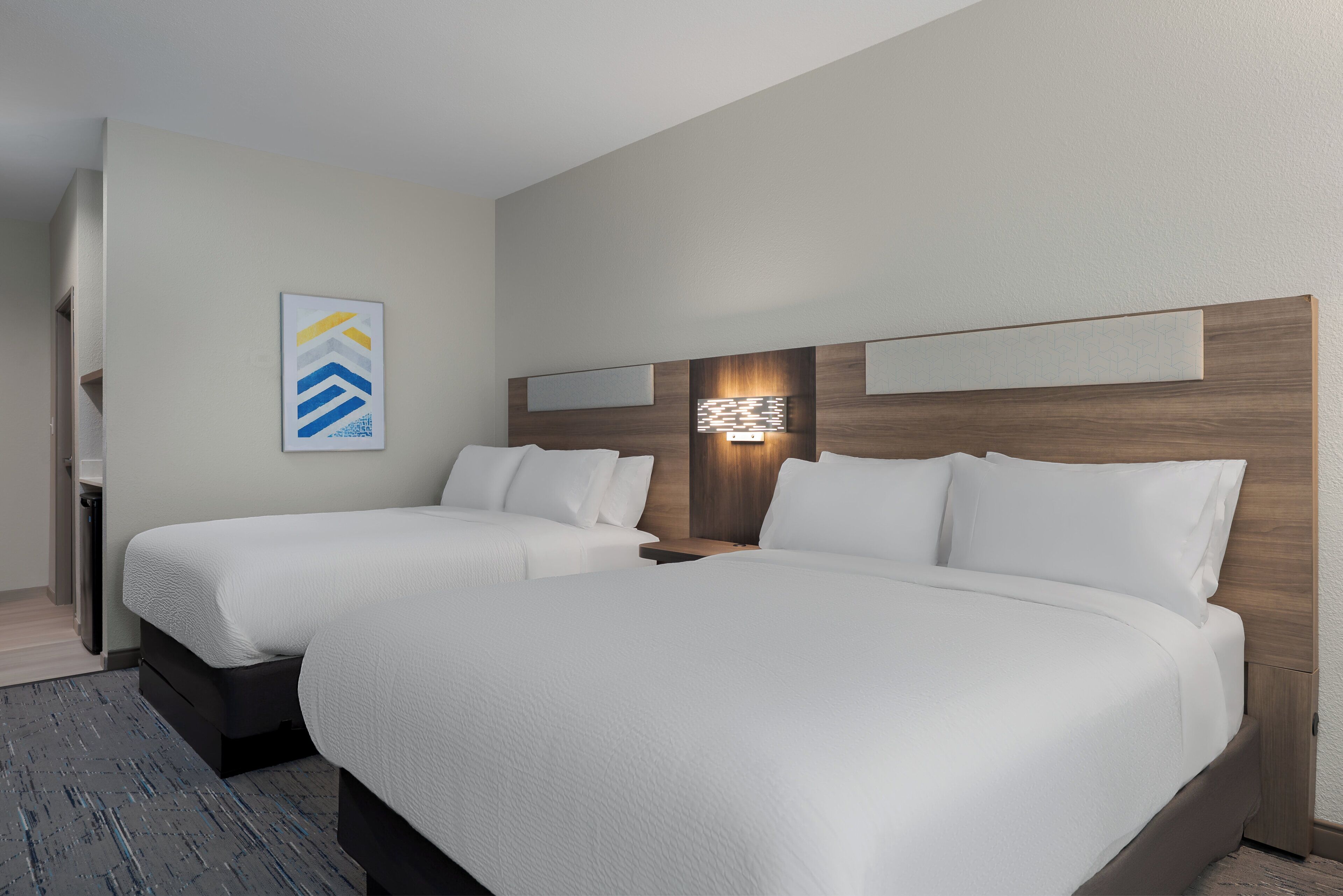 A hotel room with two beds, a headboard with built-in lighting, a wall-mounted artwork, and a small bedside table.