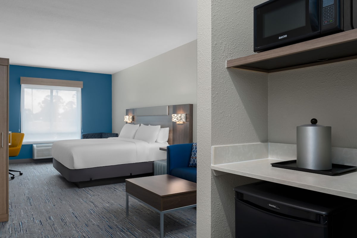A hotel room with a bed, bedside table, chair, microwave, and a small kitchen area with a microwave and a kettle.