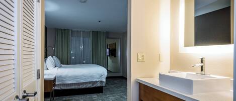 Habitació d'hotel amb un llit, un bany amb un lavabo i un mirall, i vista de l'espai a través d'una porta.