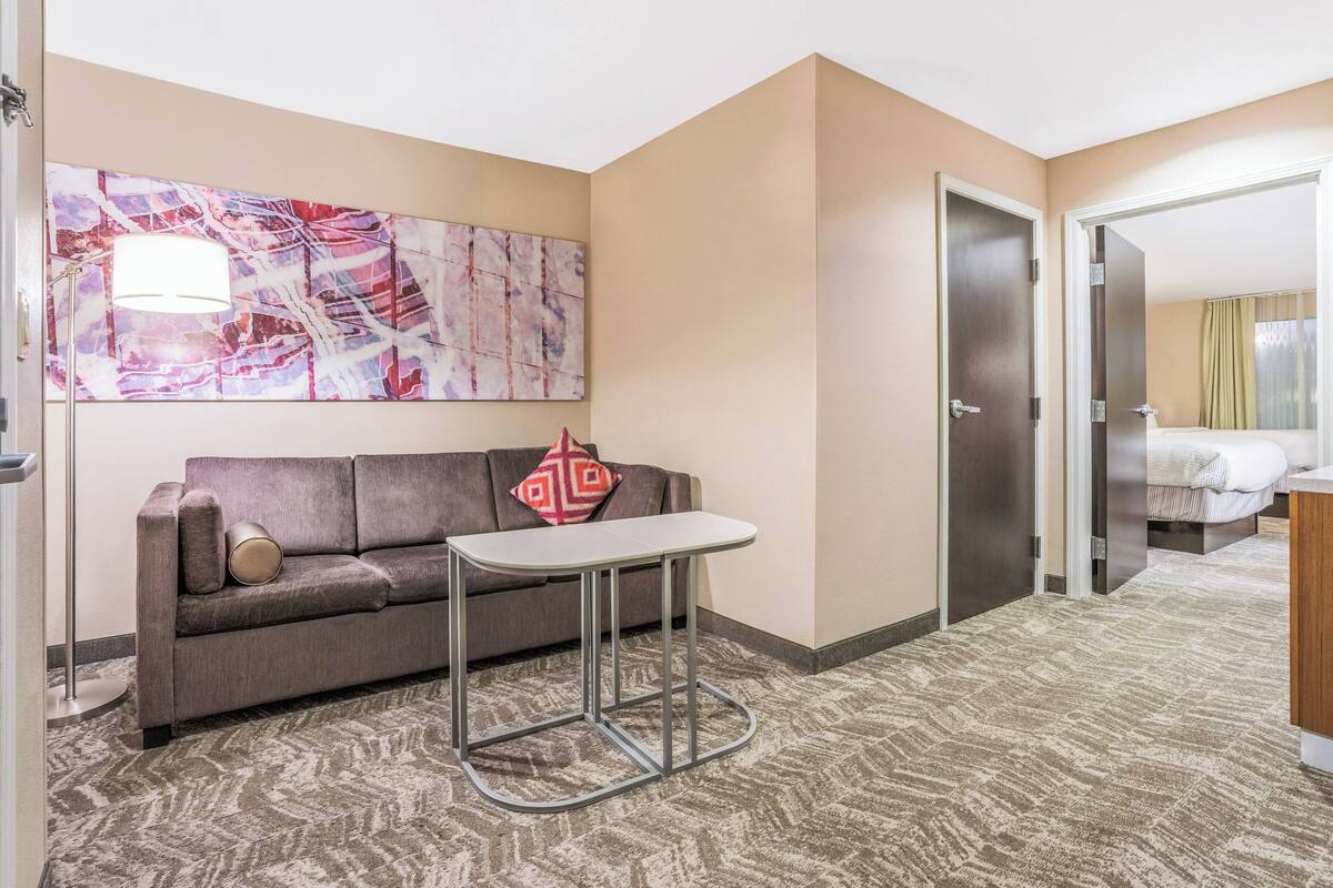A hotel room with a grey sofa, a small table, a large abstract painting on the wall, and a door leading to a bedroom.
