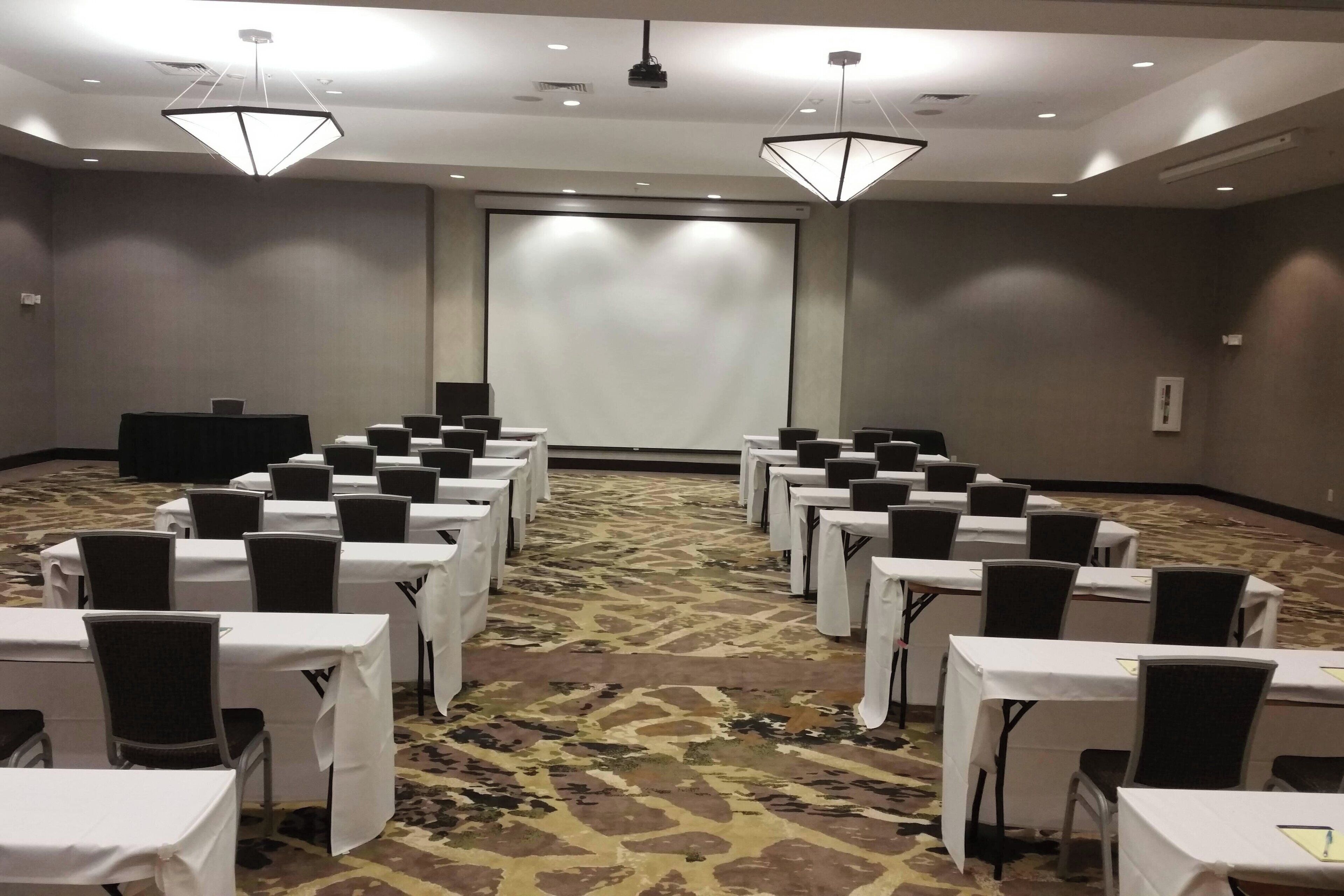 A conference room with rows of tables and chairs, a projector screen, and a carpeted floor.