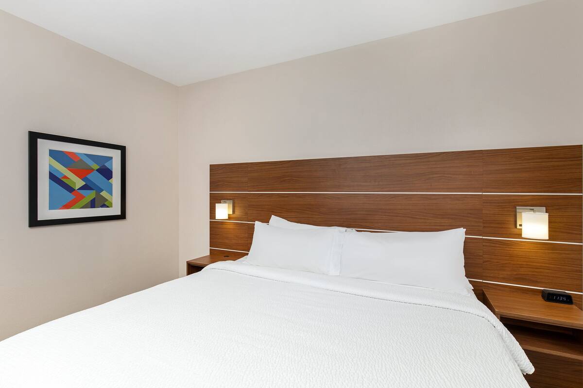 A hotel room with a large bed, wooden headboard, two bedside tables with lamps, and a framed abstract artwork on the wall.