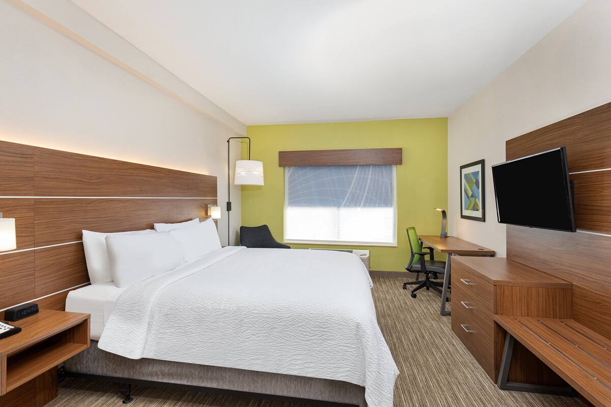 A hotel room with a large bed, bedside tables, a desk, a chair, a TV, and a window with curtains.