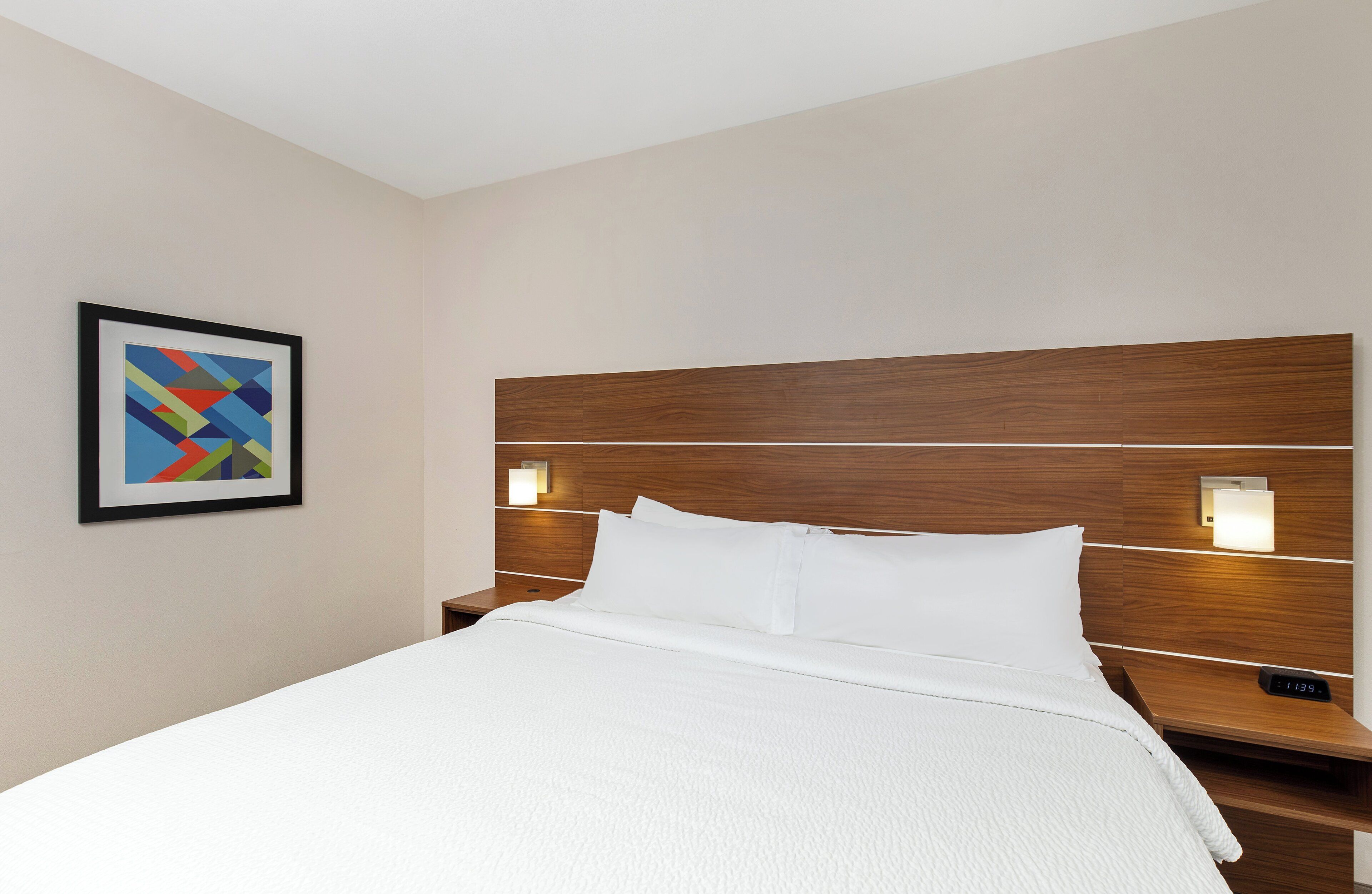 A hotel room with a bed, a nightstand, a wall-mounted lamp, and a framed picture.