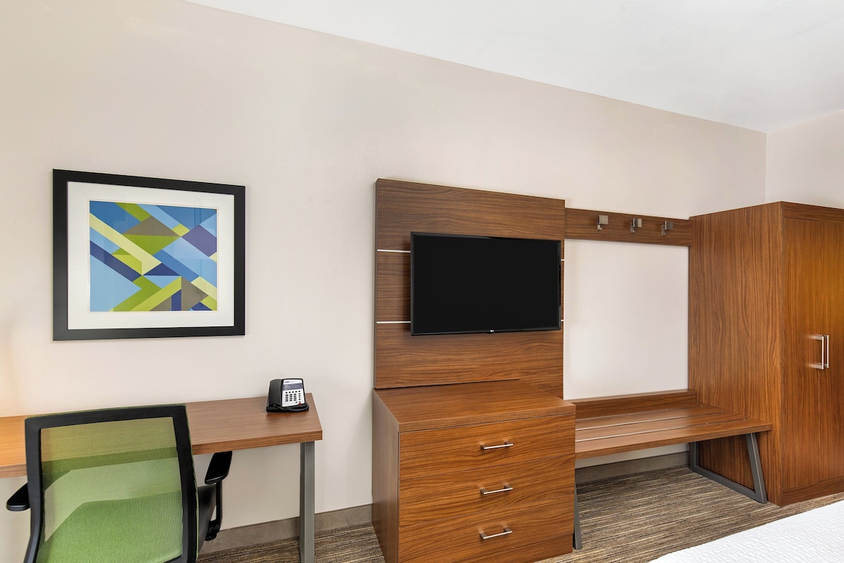 Une chambre d’hôtel avec un bureau en bois, un téléviseur fixé sur un meuble, une œuvre d’art abstraite encadrée au mur et un téléphone sur le bureau.