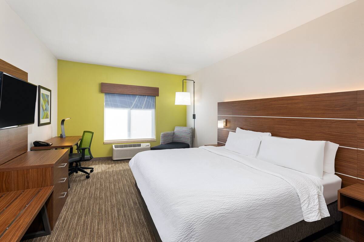 A hotel room with a large bed, a desk with a chair, a television, and a window with blinds.