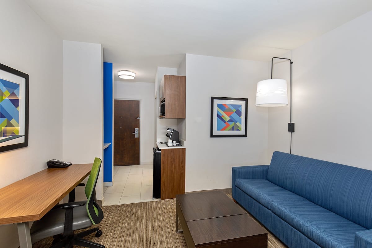 A modern hotel room with a wooden desk, a blue sofa, a small kitchen area, and a framed abstract painting.