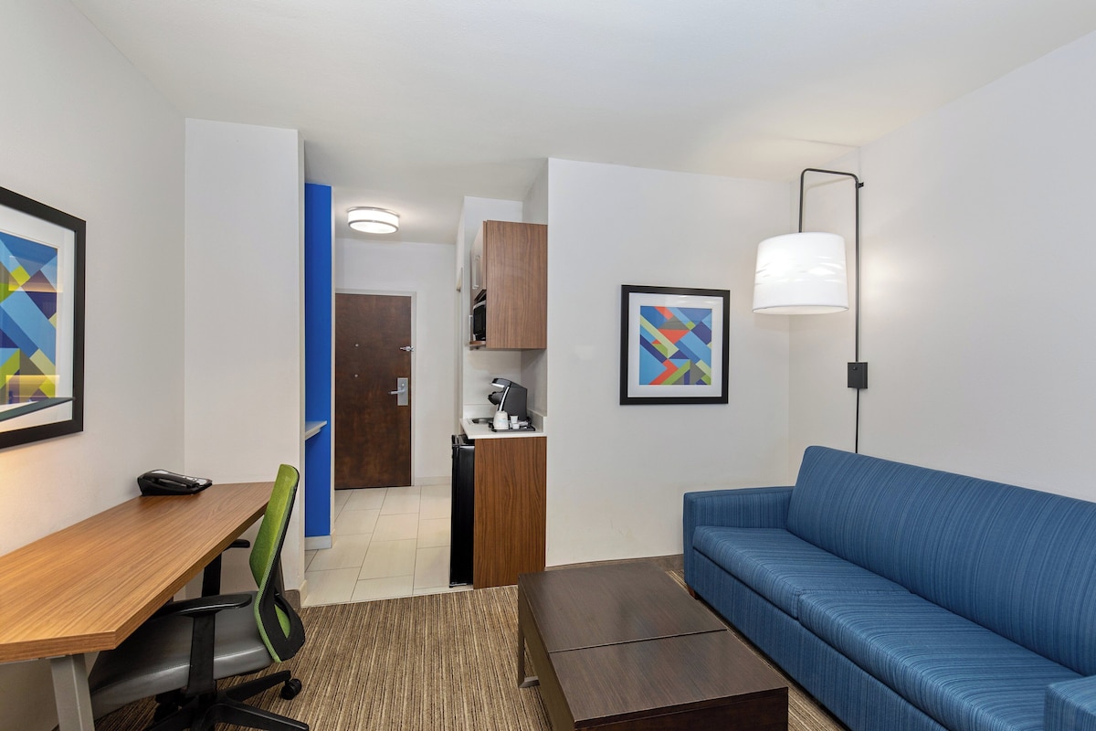 Une chambre d’hôtel moderne avec un bureau en bois, un canapé bleu, un petit coin cuisine et un tableau abstrait encadré.