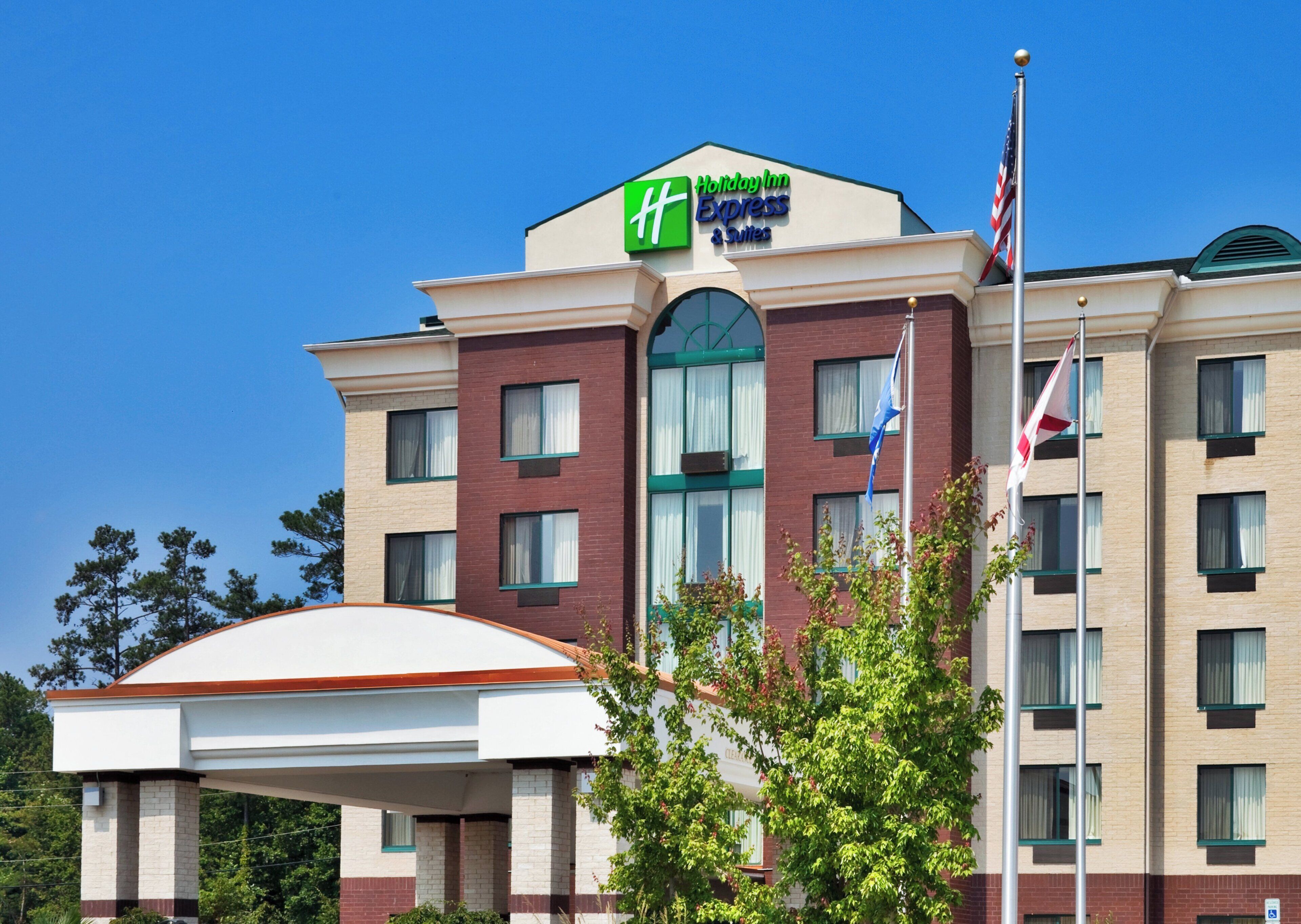 A Holiday Inn Express & Suites building with a clear blue sky, a flagpole with flags, and a covered entrance.