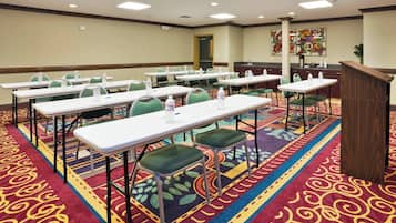 Une salle de conférence avec des rangées de tables et de chaises, équipée de bouteilles d’eau, et un pupitre à l’avant.