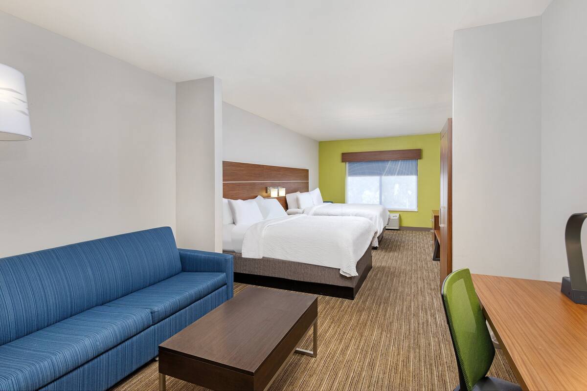A hotel room with a blue sofa, a wooden coffee table, a bed with white bedding, a green chair, and a desk with a lamp.