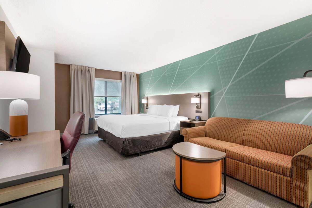 A hotel room with a bed, a sofa, a desk, a chair, a small table, and a window with curtains.