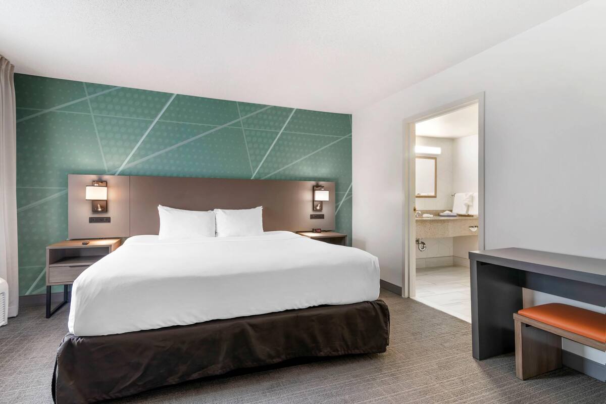 A hotel room with a large bed, bedside tables with lamps, a desk, and a bathroom visible through an open door.