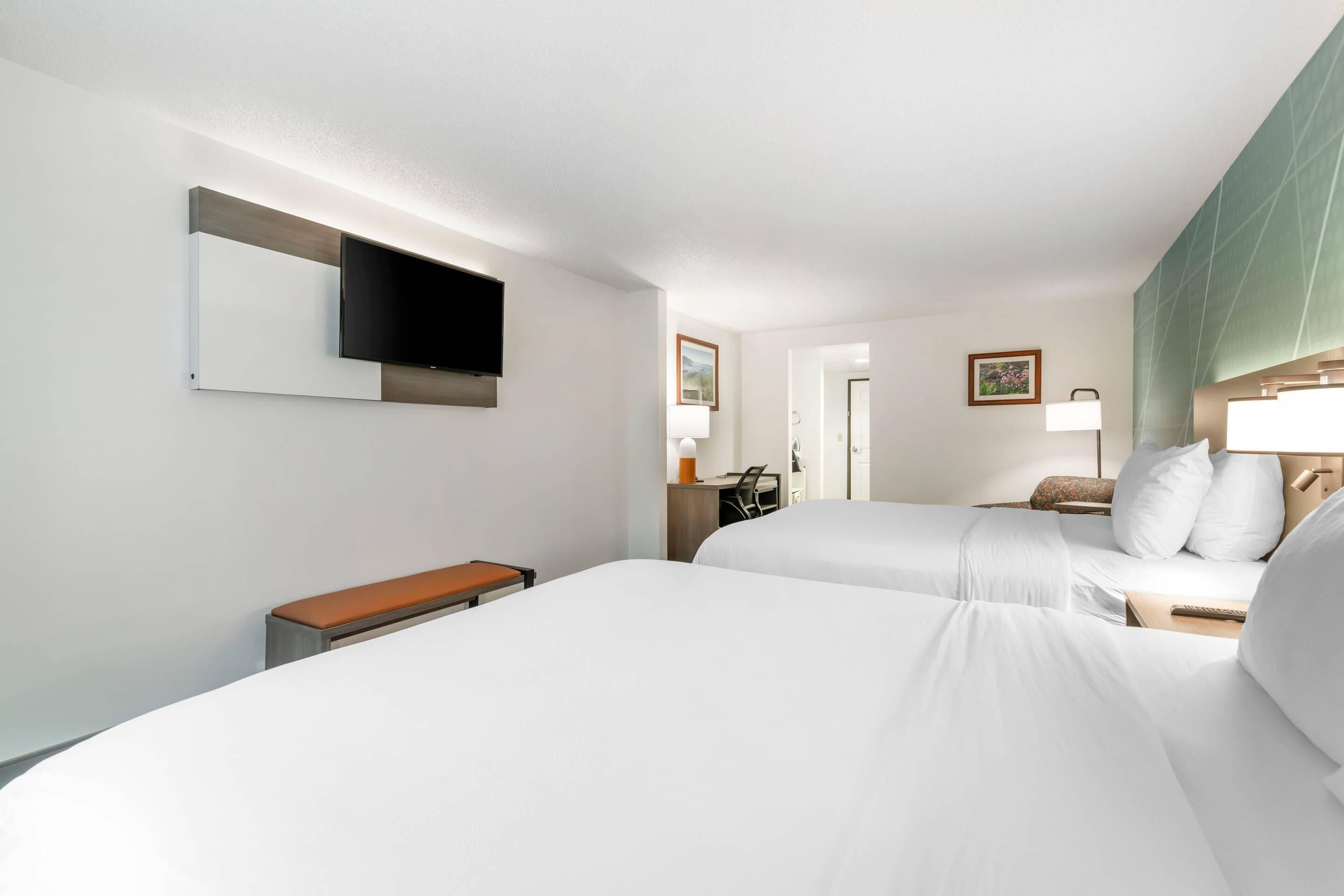 Une chambre d’hôtel avec deux lits, un bureau, une chaise, une télévision et une salle de bain visible à travers une porte ouverte.